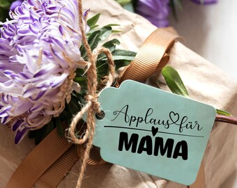 May include: A brown gift box wrapped with twine and a brown ribbon. A blue gift tag with the text "Applaus für MAMA" is attached to the box. Purple flowers are arranged on top of the box.