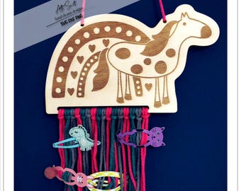Könnte beinhalten: Aus Holz ausgeschnittenes Giraffenmotiv mit einem Regenbogenbogen darüber. Die Giraffe ist braun mit weißen Flecken und hat ein rosa Herz an der Seite. Der Regenbogen ist mit weißen Punkten und rosa Herzen verziert. Der Ausschnitt hängt an einer Schnur und hat einen Fransen aus buntem Garn, der darunter hängt. Es hängen mehrere bunte Haarklammern am Garn.