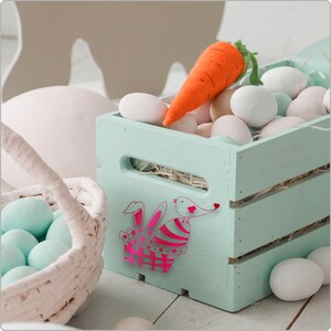 Pode incluir: Uma caixa de madeira azul claro cheia de ovos de P&aacute;scoa brancos e uma cenoura. Um desenho de coelho rosa e p&aacute;ssaro &eacute; pintado no lado da caixa.
