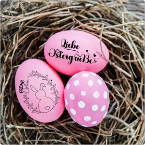 Pode incluir: Tr&ecirc;s ovos de P&aacute;scoa cor-de-rosa decorados com diferentes desenhos. Um ovo tem o texto "Liebe Ostergr&uuml;&szlig;e" em escrita cursiva preta, outro tem um coelho numa coroa com o texto "Frohe Ostern" em escrita cursiva preta, e o terceiro ovo tem bolinhas brancas.
