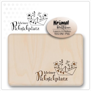 Könnte beinhalten: Ein rechteckiges Holzschneidebrett mit den Worten "kleiner Picknickplatz" und einem Papierboot-Design mit Blumen. Das Design ist in das Holz geätzt. Ein ovales Etikett lautet "Krümel Brettchen" mit zusätzlichem Text.
