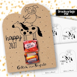 Può includere: Un cartellino in cartoncino color marrone chiaro con un disegno a linee nere di un maiale su un cavallo, con il testo "happy 2027" e "Glück zum Stapeln". Due cioccolatini incartati, uno rosso "Mon Cheri" e uno arancione "Ferrero Küsschen", sono fissati.