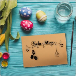 Pode incluir: Um cart&atilde;o castanho com o texto "Frohe Ostern" em escrita cursiva preta. O cart&atilde;o &eacute; decorado com uma ilustra&ccedil;&atilde;o em preto e branco de uma casa de p&aacute;ssaros pendurada numa corda com cora&ccedil;&otilde;es. H&aacute; tr&ecirc;s ovos de P&aacute;scoa numa superf&iacute;cie de madeira azul atr&aacute;s do cart&atilde;o.