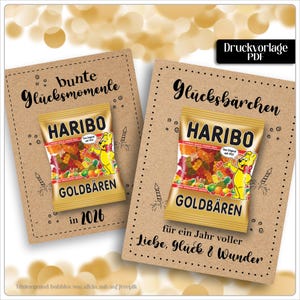 Può includere: Due biglietti di carta kraft con sacchetti di caramelle Haribo Goldbears. I biglietti hanno testo in tedesco, tra cui "bunte Glücksmomente" e "Glücksbärchen". Le borse presentano una mascotte di orsetto gommoso giallo e il testo "GOLDBÄREN".