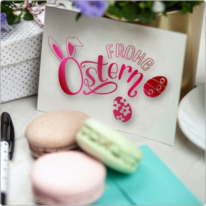 Pode incluir: Um cart&atilde;o branco com o texto "Frohe Ostern" em letras cor-de-rosa. O cart&atilde;o &eacute; decorado com um coelho rosa e ovos de P&aacute;scoa cor-de-rosa. H&aacute; tr&ecirc;s macarons sobre uma superf&iacute;cie azul em primeiro plano.
