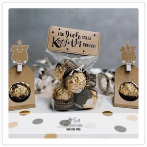 Puede incluir: Una bolsa de papel marrón con una etiqueta que dice "Para ti, debería llover confeti". La bolsa está llena de avellanas cubiertas de chocolate. La bolsa está sentada en una mesa con un recorte de cerdo marrón y blanco a cada lado.