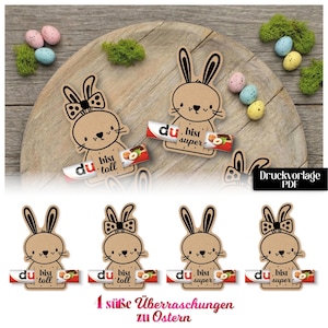 Könnte beinhalten: Osterbastelprojekt mit braunen Hasen-Ausschnitten, Schleifen und Schokoriegeln. Das Bild zeigt dekorative Eier und Moos auf einem Holztablett. Auf den Schokoriegeln steht "du bist super" und "du bist toll". Der Text am unteren Rand des Bildes lautet "1 süße Überraschungen zu Ostern".