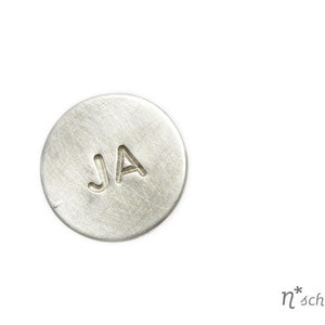 Könnte beinhalten: Ein kleiner, runder, silberfarbener Anhänger mit den Buchstaben "JA" in der Mitte. Der Anhänger hat eine gebürstete Metalloberfläche vor einem weißen Hintergrund.
