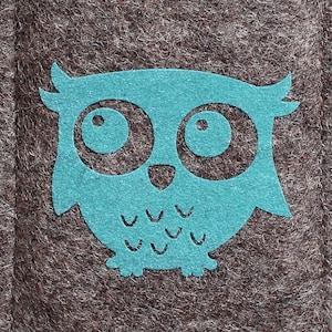 Puede incluir: Funda de teléfono de fieltro marrón con un diseño de búho turquesa. El búho está centrado en la funda y tiene ojos grandes y un contorno detallado. El nombre de la marca "ituepfelchen" está impreso en una pequeña etiqueta blanca.