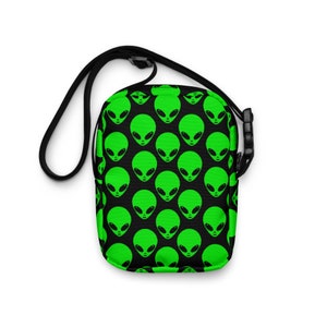 Pode incluir: Uma bolsa de ombro preta com um padrão de rosto de alienígena verde. A bolsa tem uma alça ajustável preta.