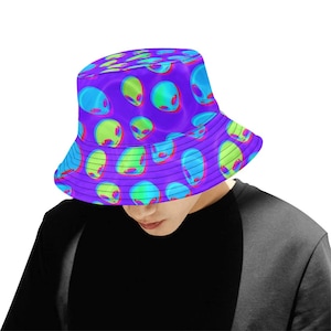 May include: A purple bucket hat with a repeating pattern of green and blue alien faces. The alien faces are in a 3D style, with a slight psychedelic effect.