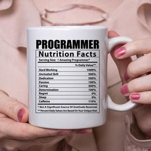 May include: White ceramic mug with a black and white graphic of a programmer nutrition facts label. The label lists various skills and attributes with a percentage daily value. The text on the mug reads "Programmer Nutrition Facts" and "Serving Size: 1 Amazing Programmer*" and "% Daily Value**". The label includes the following items: Hard Working, Unrivaled Skill, Dedication, Passion, Caring, Determination, Regret, Sleep, Caffeine. The text at the bottom of the label reads "* Not A Significant Source Of Gratitude Received." and "** Percent Daily Values Are Based On Your Unique Diet."