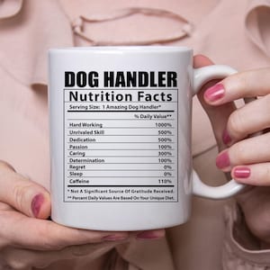 May include: White ceramic mug with a black and white graphic of a nutrition facts label for "Dog Handler". The label lists various skills and qualities with percentages, such as "Hard Working 1000%", "Unrivaled Skill 500%", "Dedication 500%", "Passion 100%", "Caring 300%", "Determination 100%", "Regret 0%", "Sleep 0%", and "Caffeine 110%". The text at the bottom of the label reads "* Not A Significant Source Of Gratitude Received. ** Percent Daily Values Are Based On Your Unique Diet."