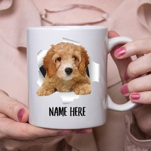 May include: White ceramic mug featuring a light brown and white puppy seemingly emerging from a torn paper effect. The text "NAME HERE" is printed in black below the puppy image. The mug has a curved handle.