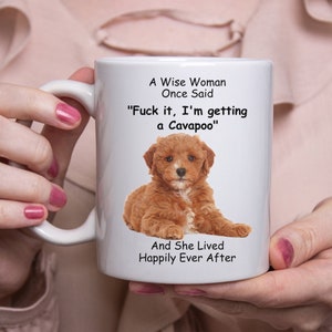May include: White ceramic mug featuring a brown Cavapoo puppy image and the text "A Wise Woman Once Said, Fuck it, I'm getting a Cavapoo, And She Lived Happily Ever After." The mug has a white handle.
