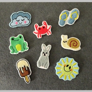Könnte beinhalten: Eine Sammlung gestickter Aufnäher mit verschiedenen Designs. Enthält eine lächelnde Wolke, eine rote Krabbe, blaue Flip-Flops, einen grünen Frosch, ein graues Kaninchen, eine Schnecke, ein Eis am Stiel und eine lächelnde Sonne, jeweils mit weißem Rand.