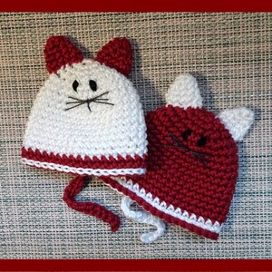 May include: Two crocheted cat hats, one white with a red trim and one red with a white trim. Both hats have black embroidered eyes and whiskers.