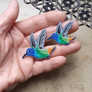 Könnte beinhalten: Zwei gestickte Kolibri-Aufnäher mit blauen, grünen und orangefarbenen Akzenten. Die Aufnäher befinden sich auf einer Hand.