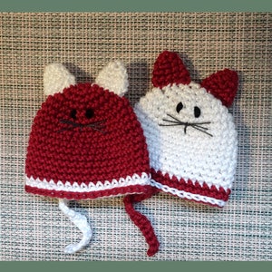 May include: Two crocheted cat hats, one red and one white, with white trim and black embroidered eyes and whiskers.