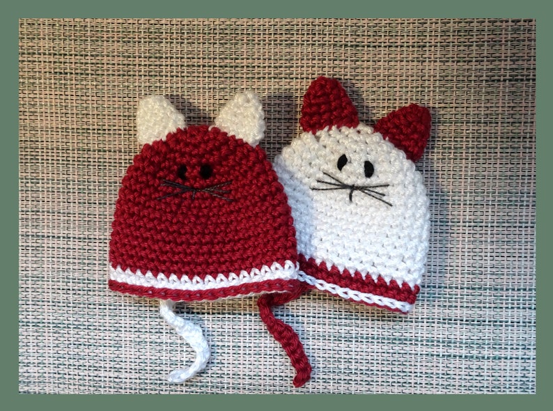 May include: Two crocheted cat hats, one red and one white, with white trim and black embroidered eyes and whiskers.