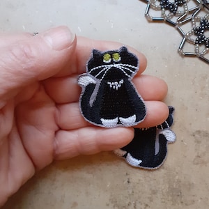 Könnte beinhalten: Zwei schwarze Filz-Katzen-Patches mit weißen Akzenten und gelben Augen. Die Katzen-Patches liegen auf einer Hand.