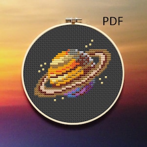 May include: A cross-stitch pattern of Saturn, a planet with rings, in a black hoop. The planet is yellow, orange, and brown with a purple and blue ring.