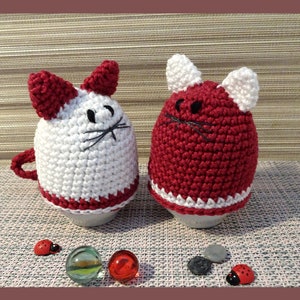 May include: Two crocheted cat egg cozies, one white with red ears and a red trim, the other red with white ears and a white trim.