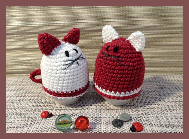 May include: Two crocheted cat egg cozies, one white with red ears and a red trim, the other red with white ears and a white trim.