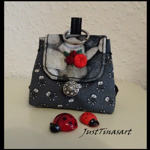 Könnte beinhalten: Eine kleine graue Stofftasche mit einem Totenkopf- und Knochenmuster. Die Tasche hat eine Klappe mit einem Blumendesign und einen silbernen Knopf. Zwei rote Marienkäfer-Figuren befinden sich vor der Tasche. Die Tasche hat einen silbernen Ring, der oben befestigt ist, und der Text "JustTinasart" ist unter der Tasche sichtbar.