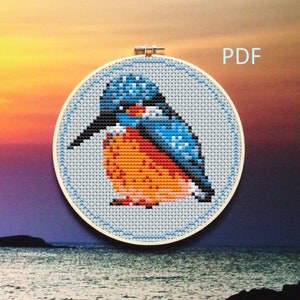 May include: A cross-stitch pattern of a blue and orange kingfisher bird on a light blue background. The pattern is in a round embroidery hoop. The image includes the word "PDF".