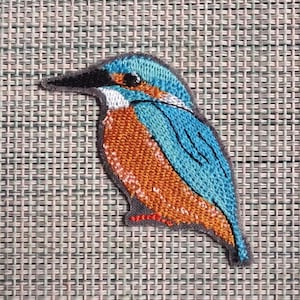 Könnte beinhalten: Gestickter Aufnäher eines Eisvogels. Der Aufnäher zeigt ein detailliertes Design mit blauem Rücken, orangefarbener Brust und schwarzem Schnabel. Der Vogel ist grau umrandet und hat ein strukturiertes Aussehen.