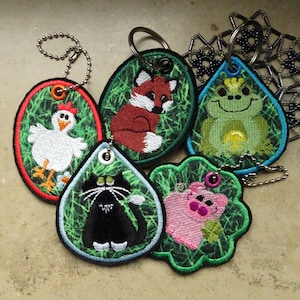 Könnte beinhalten: Fünf bunte Schlüsselanhänger mit verschiedenen Tiermotiven. Jeder Schlüsselanhänger hat einen grünen Hintergrund mit einem schwarzen Blitzmuster. Die Tiere sind ein roter Fuchs, ein grüner Frosch, ein weißes Huhn, eine schwarze Katze und ein rosa Schwein.