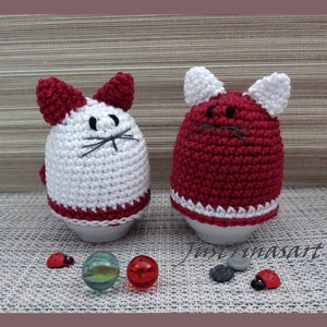 May include: Two crocheted egg cozies shaped like cats. One is white with red ears and a red bottom, the other is red with white ears and a white bottom.