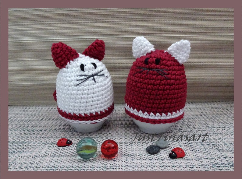 May include: Two crocheted egg cozies shaped like cats. One is white with red ears and a red bottom, the other is red with white ears and a white bottom.