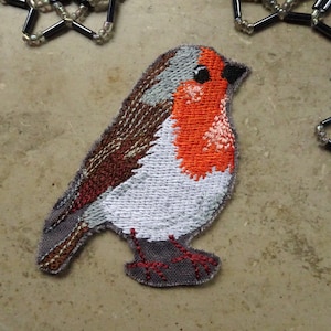 Könnte beinhalten: Ein gestickter Aufnäher eines Rotkehlchens mit roter Brust, braunen, grauen und weißen Details. Der Vogel steht auf seinen beiden Füßen mit gefalteten Flügeln.