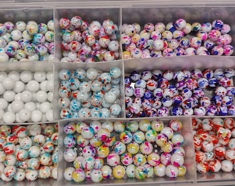 May include: A divided container filled with various round beads. Some compartments hold solid white beads, while others contain beads with colorful splattered paint designs in shades of blue, pink, purple, and yellow. The beads are likely for jewelry making.