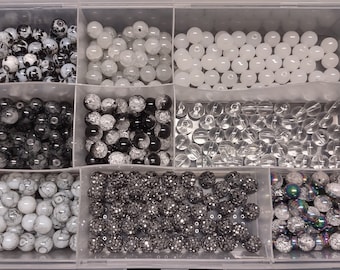 May include: A clear plastic storage box with eight compartments filled with various black and white beads. The beads are round and have different textures, including smooth, sparkly, and marbled.
