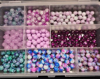 May include: A clear plastic storage box with 10 compartments filled with various colorful beads. The beads are in shades of purple, pink, blue, green, and white. Some beads are solid, while others have a marbled or speckled effect.