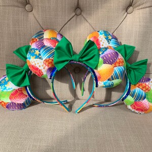 May include: A pair of colorful Easter egg-themed Minnie Mouse ears with green bows. The ears are made of fabric with a pattern of colorful eggs and flowers.