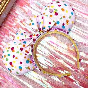 May include: Pink Minnie Mouse ears with a confetti pattern and a gold headband. The bow is made of pink fabric with colorful confetti.