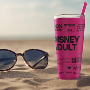 Puede incluir: Un vaso rosa con un diseño en blanco y negro que dice "100% Inspirado, Libre de Estrés, Disney Adulto". El vaso está en una playa de arena con un par de gafas de sol marrones.