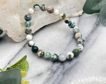 May include: A beaded bracelet featuring round, natural stone beads in shades of white, green, and brown. The bracelet is strung on an elastic cord and is displayed on a marble surface.