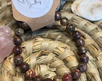 May include: A beaded bracelet featuring round, speckled beads in shades of brown, red, and cream. The bracelet is displayed on a woven straw mat, with a tag that reads "LUNAR NEPTUNE Jewellery".