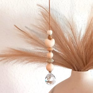 Könnte beinhalten: Dekorativer Hängeornament mit Holzperlen, einer herzförmigen Perle und einem Kristallanhänger. Das Ornament hängt vor einer Vase mit getrocknetem Pampasgras und schafft eine neutrale, böhmische Ästhetik.