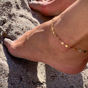 Könnte beinhalten: Goldenes Fußkettchen mit kleinen runden Anhängern an einer zarten Kette. Das Fußkettchen wird an einem Fußgelenk getragen, wobei der Fuß auf Sand ruht.