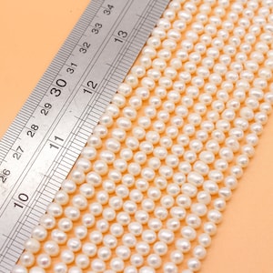 Puede incluir: Una hebra de perlas de agua dulce blancas, que mide aproximadamente 28 cm de largo, se coloca junto a una regla. Las perlas son uniformes en tamaño y forma, con un acabado liso y brillante.