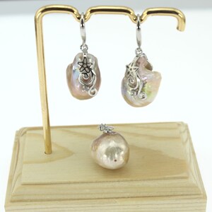 May include: A set of pearl earrings and a pendant necklace with silver floral accents. The pearls are a soft pink color and have an iridescent sheen. The earrings and pendant are displayed on a wooden stand.