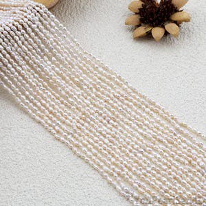 May include: Strands of small, off-white freshwater pearls arranged diagonally across a textured surface. The pearls have an irregular shape and a subtle luster. A dried flower is in the upper right corner.