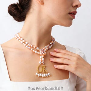 Peut inclure: Un collier à double rang composé de perles d'eau douce ovales, blanches et rose pâle irisées. Le collier présente un pendentif en métal doré avec les lettres OK, agrémenté de petites perles blanches suspendues sous les lettres.