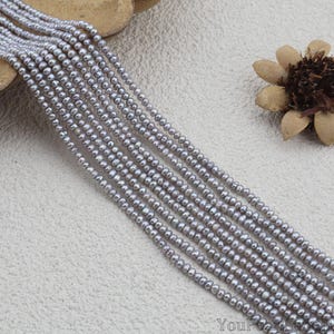 May include: Strands of small, gray pearls arranged diagonally across the frame. The pearls have a subtle luster and are uniform in size. A dried flower is visible in the upper right corner. The background is a textured, off-white surface.